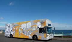 An art bus decorated with black and white illustrations, parked beside a seaside road under a clear blue sky.