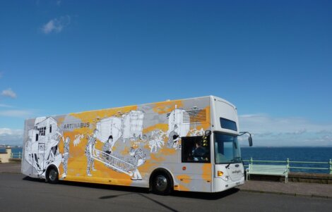 An art bus decorated with black and white illustrations, parked beside a seaside road under a clear blue sky.