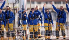 A group of jubilant sailors in matching blue jackets and yellow pants wave enthusiastically aboard a sailboat.