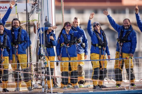 A group of jubilant sailors in matching blue jackets and yellow pants wave enthusiastically aboard a sailboat.