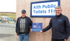 Two men standing beside a sign that reads "aith public toilets," one of them holding a small award that says "coastal aquaculture scotland.