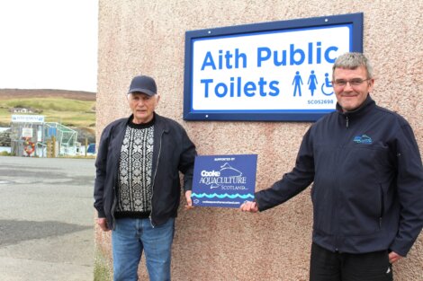 Two men standing beside a sign that reads "aith public toilets," one of them holding a small award that says "coastal aquaculture scotland.