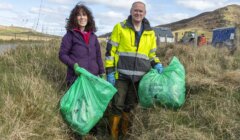 Two adults, a woman and a man, holding green trash bags during a clean-up event in a grassy field with hills and buildings in the background.