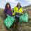 Two adults, a woman and a man, holding green trash bags during a clean-up event in a grassy field with hills and buildings in the background.