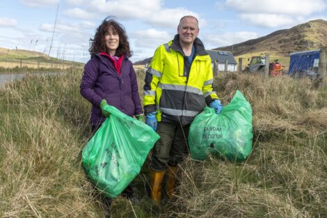 Two adults, a woman and a man, holding green trash bags during a clean-up event in a grassy field with hills and buildings in the background.