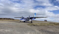 A small blue and white aircraft with "shetland.gov.uk" on the side is parked on a gravel surface under a partly cloudy sky.