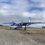 A small blue and white aircraft with "shetland.gov.uk" on the side is parked on a gravel surface under a partly cloudy sky.