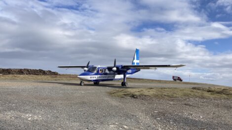 A small blue and white aircraft with "shetland.gov.uk" on the side is parked on a gravel surface under a partly cloudy sky.