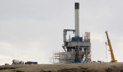 Industrial construction site with a large crane and a partially built structure featuring a tall chimney, surrounded by machinery and building materials under a cloudy sky.