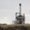 Industrial construction site with a large crane and a partially built structure featuring a tall chimney, surrounded by machinery and building materials under a cloudy sky.