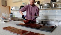 A man pouring melted chocolate into molds on a kitchen counter surrounded by cooking utensils and containers.