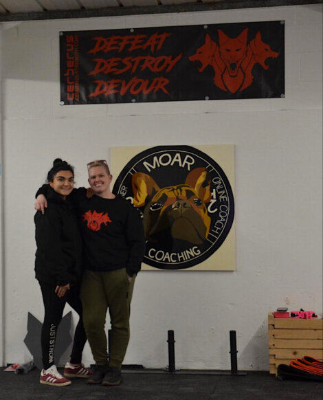 Two people smiling and posing in a gym, standing in front of banners and a large sign featuring a bulldog.