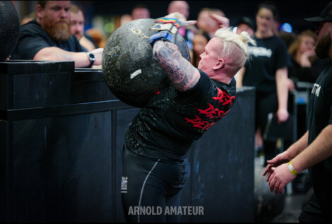 A strongman competitor lifts a heavy atlas stone during a competitive event, surrounded by spectators and judges.