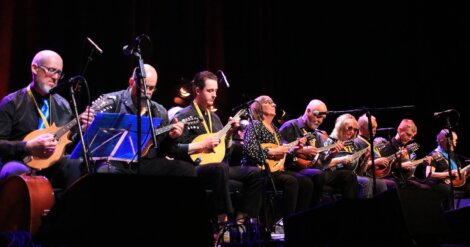 An ensemble of seven musicians playing mandolins on stage under soft blue and orange lighting.