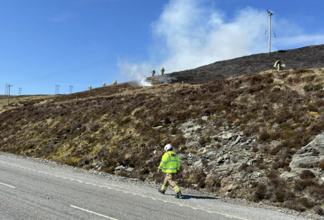 Firefighters and emergency personnel tackle a hillside fire near a road under a clear blue sky.