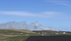 Smoke rises from a hill behind a rural landscape with houses and wind turbines under a clear blue sky.