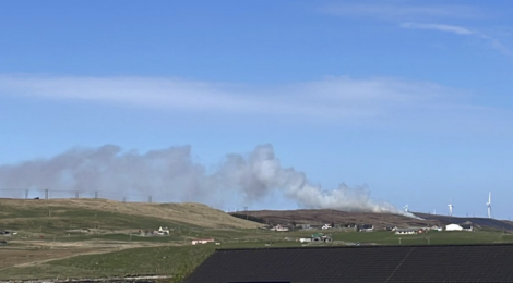 Smoke rises from a hill behind a rural landscape with houses and wind turbines under a clear blue sky.