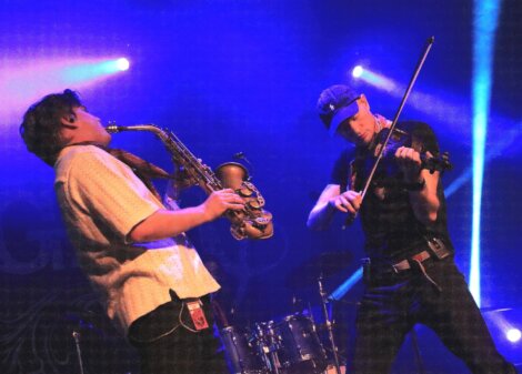 Two musicians on stage, one playing a saxophone and the other a violin, under blue stage lighting.