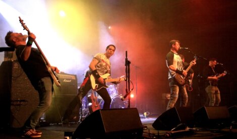 Four band members performing on stage with guitars under colorful stage lights.