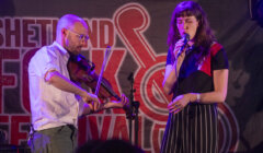 Two musicians perform on stage, a man playing a violin and a woman singing into a microphone, with a banner reading "shetland folk festival" in the background.