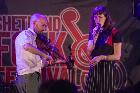 Two musicians perform on stage, a man playing a violin and a woman singing into a microphone, with a banner reading "shetland folk festival" in the background.