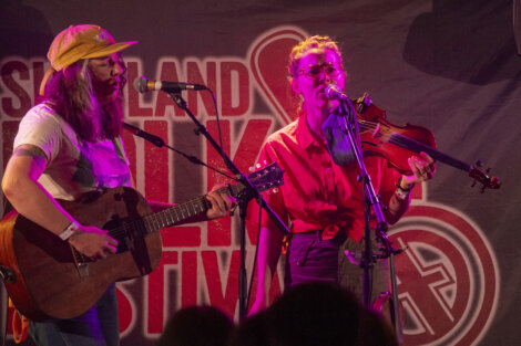 Two musicians performing on stage, one playing a guitar and the other a violin, both singing into microphones, with a festival logo in the background.