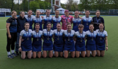 Women's field hockey team posing with medals on a sports field, wearing blue uniforms and smiles, with a coach on the left.