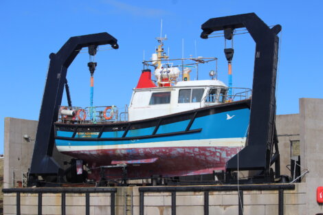 A blue and white boat hoisted by a large black boat lift against a clear blue sky.