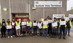 Group of workers in safety vests holding "unite" signs in front of sullom voe terminal entrance.