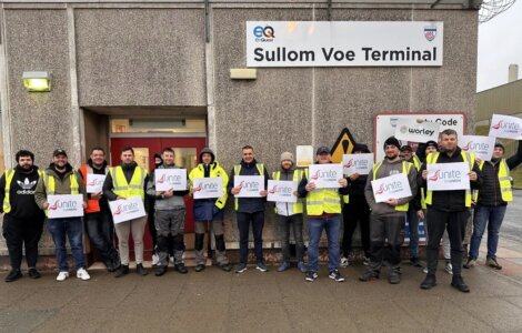 Group of workers in safety vests holding "unite" signs in front of sullom voe terminal entrance.