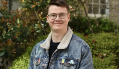 A young man wearing glasses and a denim jacket adorned with patches smiles in a garden setting.