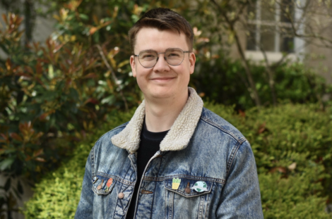 A young man wearing glasses and a denim jacket adorned with patches smiles in a garden setting.