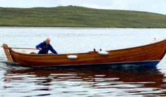 A man in a blue wetsuit rows a wooden boat on a calm sea, with a hilly coastline in the background.