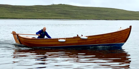 A man in a blue wetsuit rows a wooden boat on a calm sea, with a hilly coastline in the background.