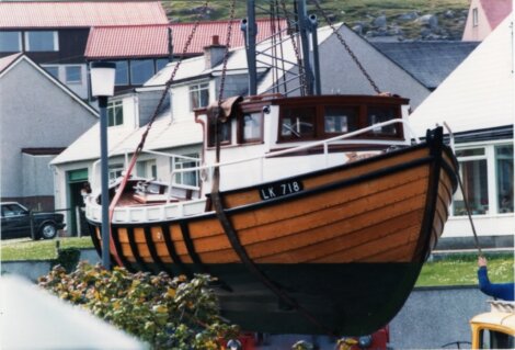 A wooden fishing boat with registration "LK 718," displayed on land against a backdrop of residential houses and greenery.