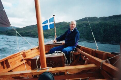 A man in a blue jacket sits at the helm of a wooden sailboat, flying a Finnish flag, on a lake with hilly landscape in the background.