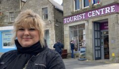 Woman smiling in front of a tourist center with stone buildings and a man in the background.