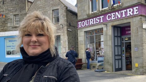 Woman smiling in front of a tourist center with stone buildings and a man in the background.