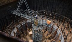 Aerial view of a construction worker on a crane inside a large, circular concrete formwork filled with scaffolding.