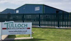 A green and white "Tulloch Developments" sign in front of a blue industrial building with a green fence under a clear sky.