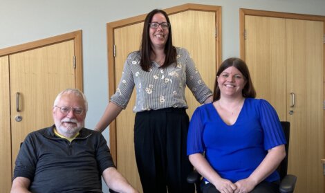 Three people are posing for a photo in an office setting. The man is seated on the left, a standing woman is in the middle, and a seated woman is on the right. They are all smiling.