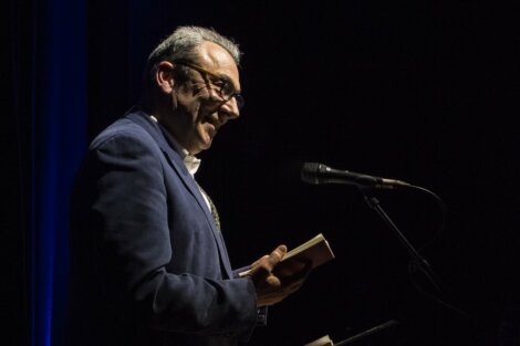 A man in a blue suit and glasses reads from a book while speaking into a microphone on a dark stage.