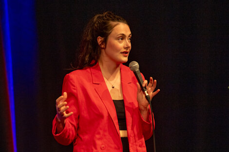 Person in a red blazer speaking into a microphone on a dark stage, gesturing with their hands.