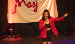 A woman in a red blazer and black outfit performs on stage with a microphone, standing in front of a white banner with red text.