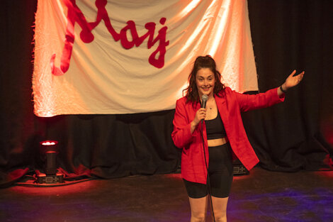 A woman in a red blazer and black outfit performs on stage with a microphone, standing in front of a white banner with red text.