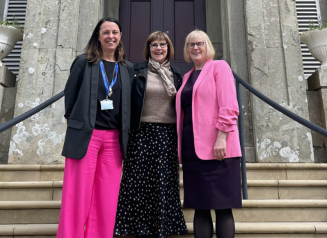 Three women stand on stairs in front of a building. The woman on the left wears a black blazer and pink pants, the woman in the middle wears a black and white polka dot skirt, and the woman on the right wears a pink blazer.