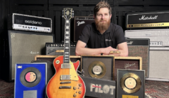 A man with a beard is sitting behind a collection of guitar amplifiers, a vintage electric guitar, and several framed gold records.