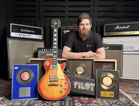 A man with a beard is sitting behind a collection of guitar amplifiers, a vintage electric guitar, and several framed gold records.