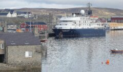 A large ship docked at a harbor with stone buildings and smaller boats in the water; hilly landscape in the background.