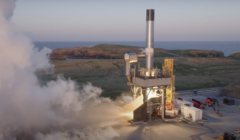 A rocket stands on a launch pad emitting smoke and preparing for liftoff, with a coastal landscape and ocean in the background.